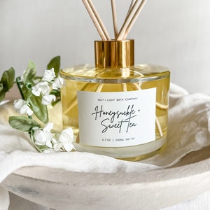 May include: A clear glass diffuser bottle with a gold lid and wooden sticks. The bottle contains a yellow liquid and has a white label that reads "Salt + Light Bath Company" and "Honeysuckle + Sweet Tea" with the text "6.7 oz | 200ml NET WT".