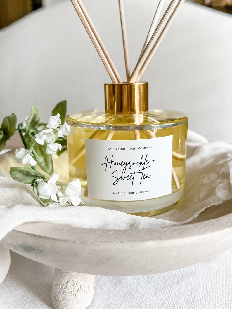 May include: A clear glass diffuser bottle with a gold lid and wooden sticks. The bottle contains a yellow liquid and has a white label that reads "Salt + Light Bath Company" and "Honeysuckle + Sweet Tea" with the text "6.7 oz | 200ml NET WT".