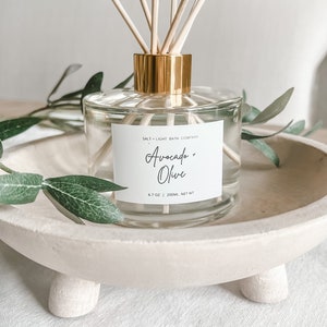 K&ouml;nnte beinhalten: Ein klarer Glasdiffusor mit goldfarbenem Kragen und Holzst&auml;bchen. Das Etikett lautet "Avocado + Olive" und "Salt + Light Bath Company". Der Diffusor steht in einer cremefarbenen Holzschale, mit gr&uuml;nen Bl&auml;ttern.