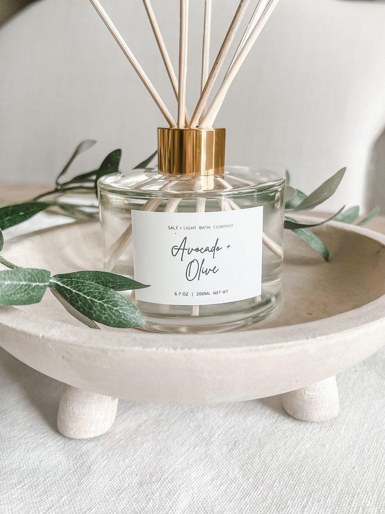 K&ouml;nnte beinhalten: Ein klarer Glasdiffusor mit goldfarbenem Kragen und Holzst&auml;bchen. Das Etikett lautet "Avocado + Olive" und "Salt + Light Bath Company". Der Diffusor steht in einer cremefarbenen Holzschale, mit gr&uuml;nen Bl&auml;ttern.