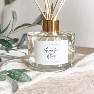 K&ouml;nnte beinhalten: Ein klarer Glasdiffusor mit goldfarbenem Kragen und Holzst&auml;bchen. Das Etikett lautet "Avocado + Olive" und "6.7 OZ | 200ml NET WT". Der Diffusor steht auf einem wei&szlig;en Tuch mit gr&uuml;nen Bl&auml;ttern im Hintergrund.
