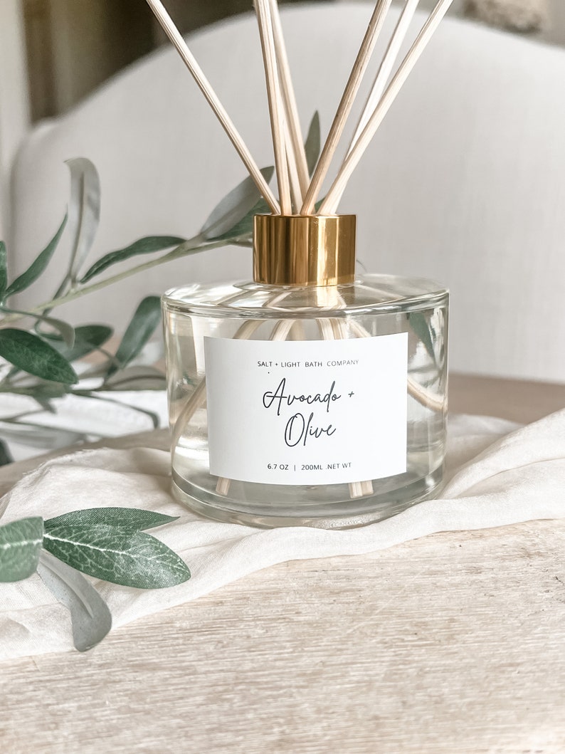 K&ouml;nnte beinhalten: Ein klarer Glasdiffusor mit goldfarbenem Kragen und Holzst&auml;bchen. Das Etikett lautet "Avocado + Olive" und "6.7 OZ | 200ml NET WT". Der Diffusor steht auf einem wei&szlig;en Tuch mit gr&uuml;nen Bl&auml;ttern im Hintergrund.