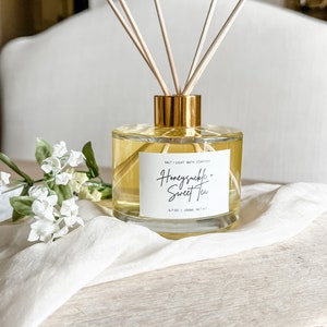 May include: A clear glass diffuser bottle with a gold lid and wooden sticks. The bottle is filled with a yellow liquid and has a label that reads "Honeysuckle + Sweet Tea" and "Salt + Light Bath Company". The bottle is sitting on a wooden surface.