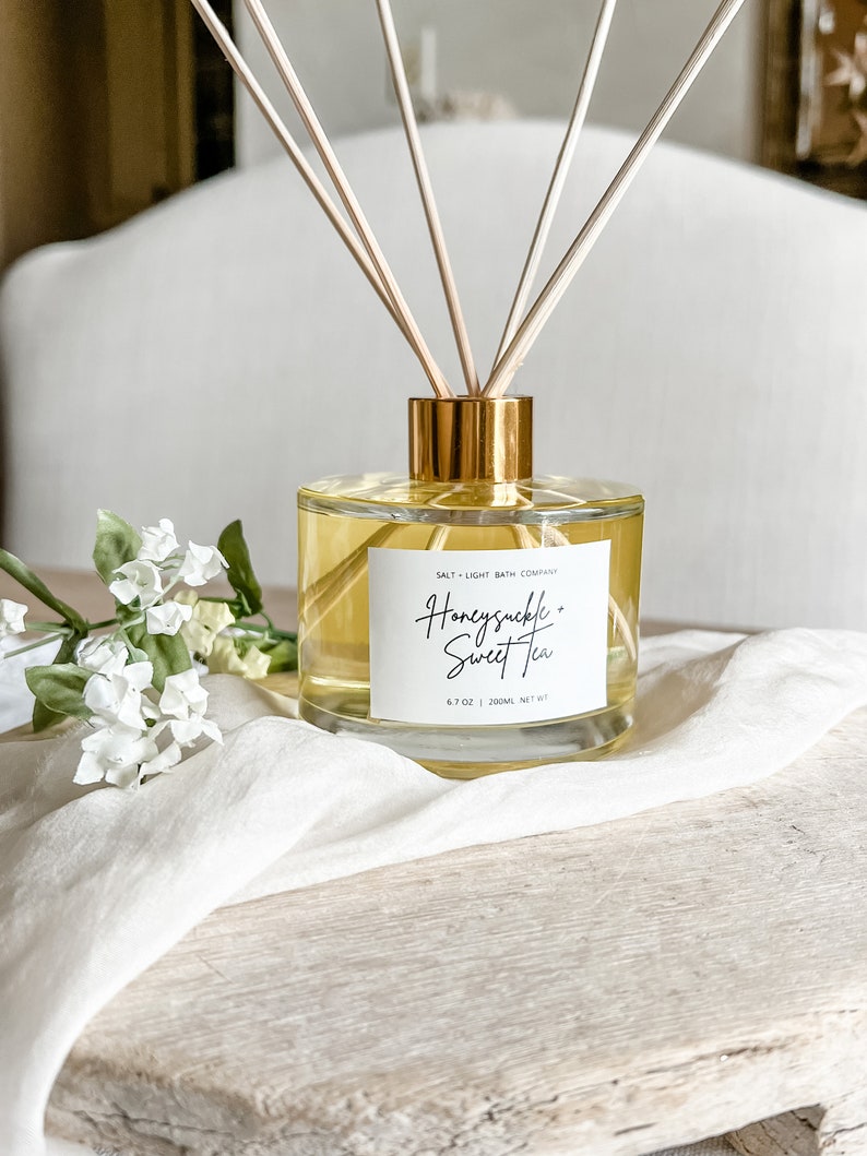 May include: A clear glass diffuser bottle with a gold lid and wooden sticks. The bottle is filled with a yellow liquid and has a label that reads "Honeysuckle + Sweet Tea" and "Salt + Light Bath Company". The bottle is sitting on a wooden surface.