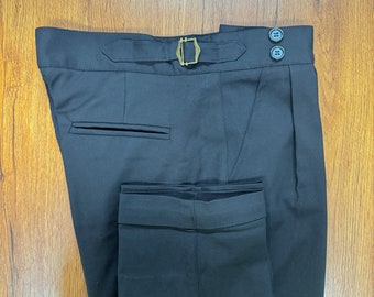 May include: Black dress pants with a tailored fit. The pants feature a buckle closure, two buttons, and a pocket. The fabric appears to be a smooth, solid color. The pants are folded neatly on a wooden surface.