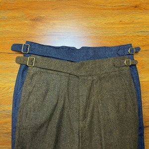 Puede incluir: Un pantalón de tweed marrón y azul con un patrón de espiga. El pantalón tiene dos correas ajustables con hebillas de latón.