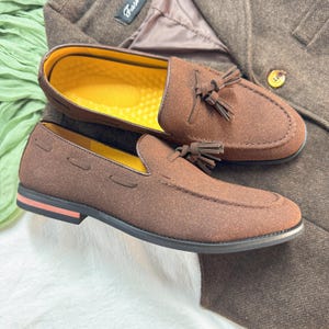 May include: A pair of brown suede tassel loafers with a yellow insole. The loafers have a black sole and a pink stripe on the bottom.