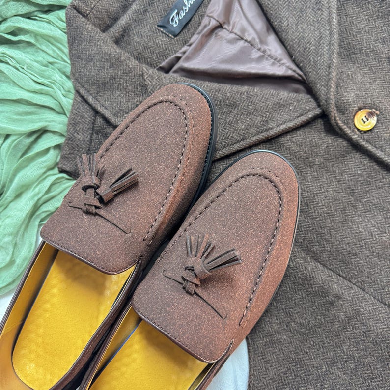 May include: A pair of brown suede loafers with tassels. The loafers have a yellow insole and are shown on a brown tweed fabric.