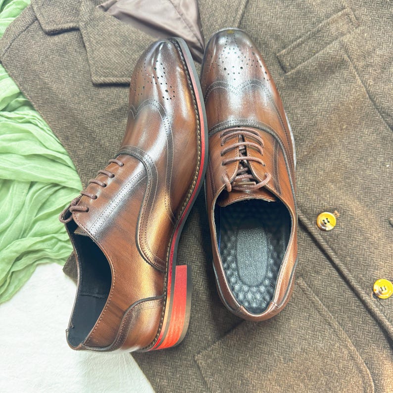 May include: A pair of brown leather lace-up shoes with wingtip detailing. The shoes have a red trim along the sole and are on a brown tweed fabric background.