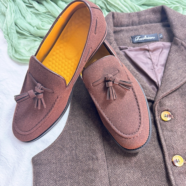 May include: A pair of brown suede tassel loafers with a yellow insole. The loafers are on a brown tweed fabric background.