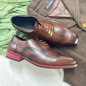 May include: A pair of brown leather lace-up shoes with a red trim on the sole. The shoes have a wingtip design and are made of a distressed leather.
