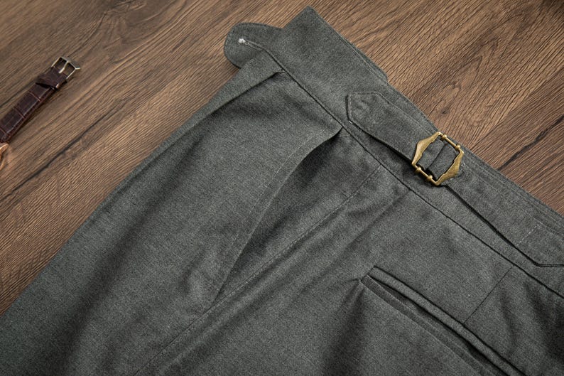 K&ouml;nnte beinhalten: Nahaufnahme einer dunkelgrauen Hose mit einer einzigen vorderen Tasche und einer Messing-Schnalle am Bund.