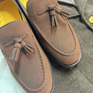 May include: A pair of brown suede loafers with tassels. The loafers have a pointed toe and a stitched seam around the edge.