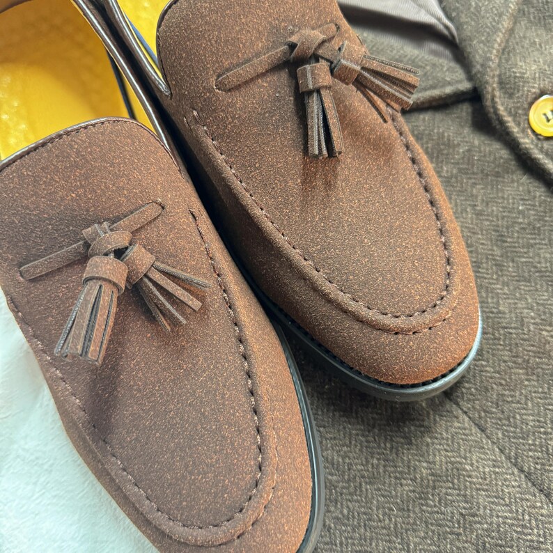 May include: A pair of brown suede loafers with tassels. The loafers have a pointed toe and a stitched seam around the edge.