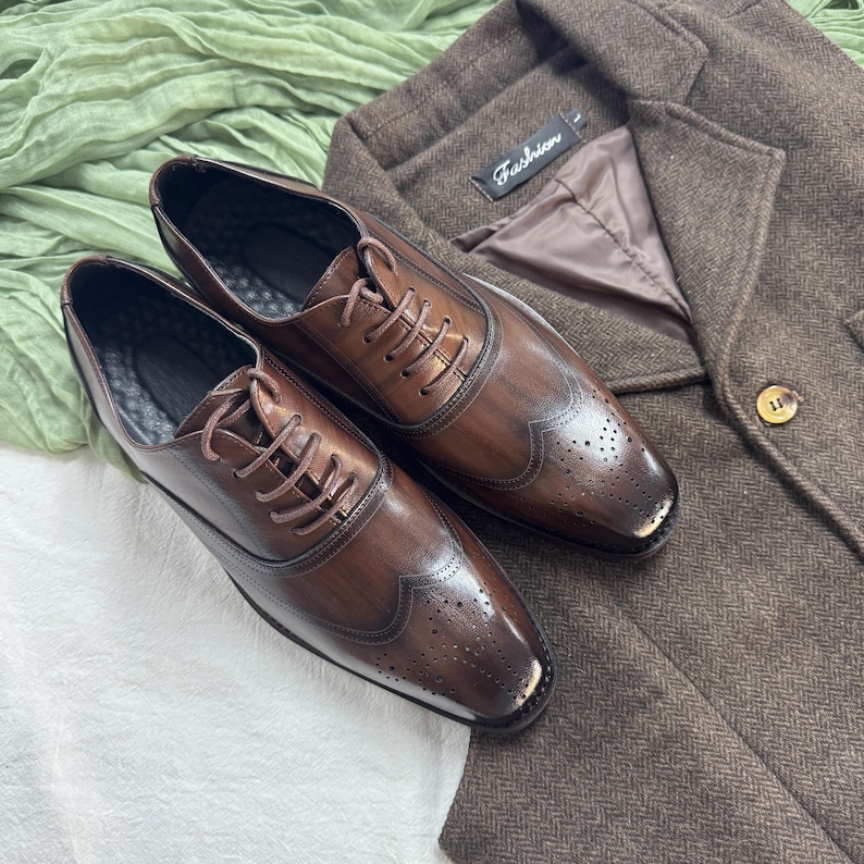 May include: A pair of brown leather lace-up shoes with wingtip detailing. The shoes have a distressed look and are sitting on a brown fabric surface.