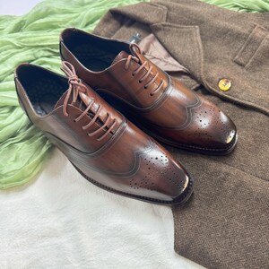 May include: Two brown leather lace-up shoes with wingtip detailing. The shoes have a brogue pattern and are on a white surface.