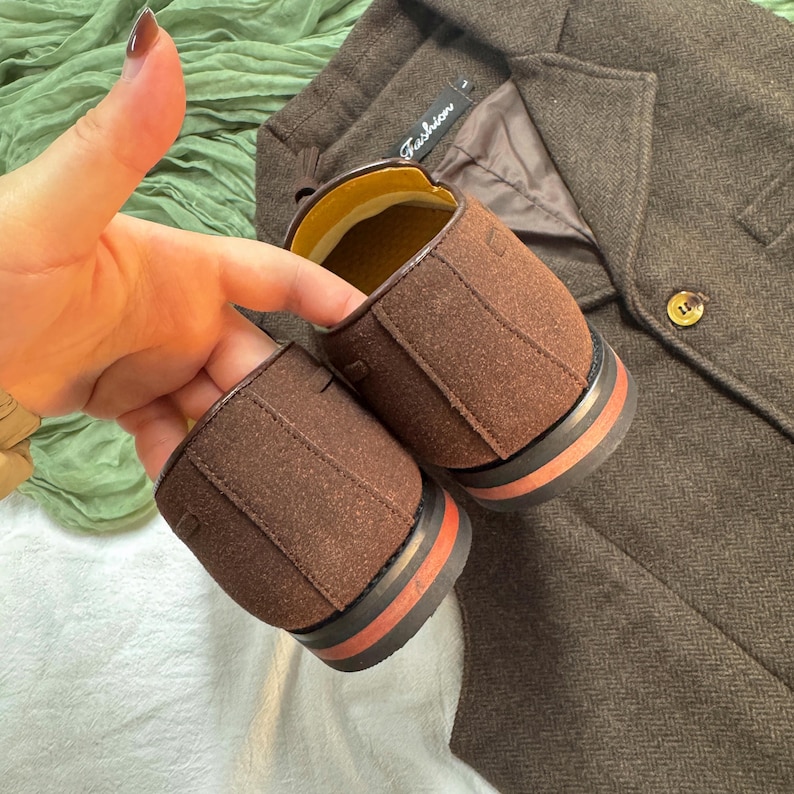 May include: A pair of brown suede shoes with red and black trim. The shoes are on a brown tweed fabric.