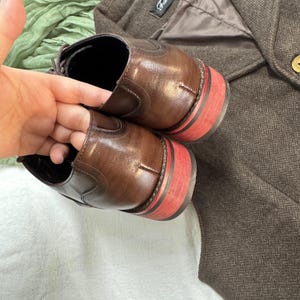 May include: A pair of brown leather shoes with red trim on the soles. The shoes are laced and have a classic design.