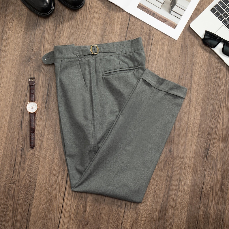 K&ouml;nnte beinhalten: Eine graue Hose mit einem G&uuml;rtelschlaufen und einem einzigen Knopfverschluss. Die Hose ist in der H&auml;lfte gefaltet und liegt flach auf einer Holzoberfl&auml;che.