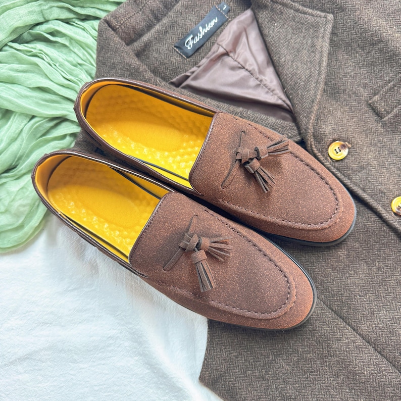 May include: A pair of brown suede tassel loafers with yellow insoles. The loafers are on a white surface with a brown fabric in the background.