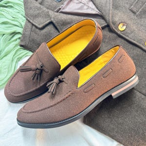 May include: A pair of brown suede tassel loafers with yellow insoles. The loafers have a brown leather sole and a tassel detail on the front.