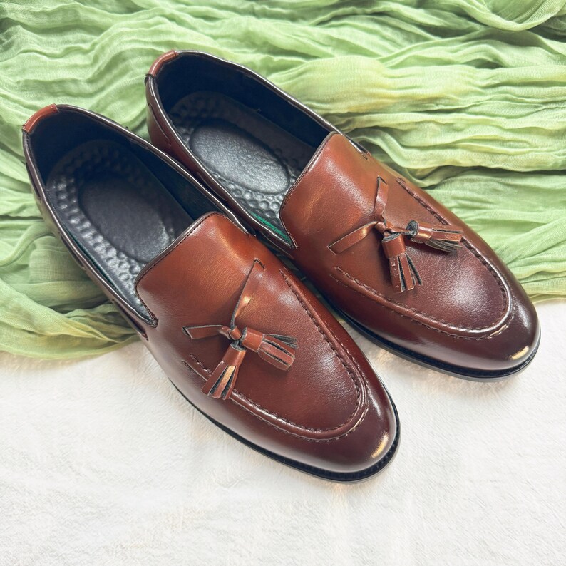 May include: A pair of brown leather tassel loafers with a slip-on design. The loafers have a dark brown tassel on each shoe.