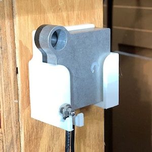 Puede incluir: Un soporte de pared blanco impreso en 3D con una placa de metal plateada unida a él. El soporte tiene un agujero para que pase un cable. El soporte está unido a una superficie de madera.