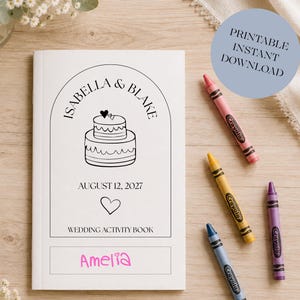 Op de afbeelding: Een wit trouwactiviteitenboek met een zwarte lijntekening van een taart met twee lagen, harten en de namen "Isabella & Blake". De datum "12 augustus 2027" staat onder de taart gedrukt. De naam "Amelia" is in roze geschreven. Crayola-kleurpotloden omringen het boek.