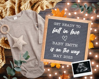 May include: A grey letter board with the text "GET READY TO fall in love ♡ BABY SMITH is on the way MAY 2023". The board is surrounded by autumnal decorations, including a baby onesie, a star-shaped toy, and a eucalyptus branch.