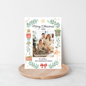 K&ouml;nnte beinhalten: Eine Weihnachtskarte mit einer Aquarell-Illustration eines Nussknackers, einer Ballerina und einer Geschenkbox. Die Karte zeigt ein Familienfoto eines Paares und ihres Kindes. Der Text auf der Karte lautet "Frohe Weihnachten 2024" und "mit Liebe, DIE FAMILIE ANDERSON".