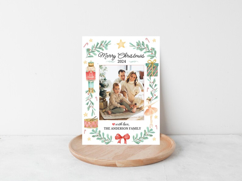 K&ouml;nnte beinhalten: Eine Weihnachtskarte mit einer Aquarell-Illustration eines Nussknackers, einer Ballerina und einer Geschenkbox. Die Karte zeigt ein Familienfoto eines Paares und ihres Kindes. Der Text auf der Karte lautet "Frohe Weihnachten 2024" und "mit Liebe, DIE FAMILIE ANDERSON".