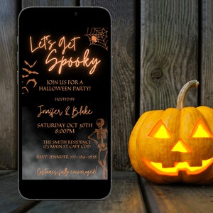 K&ouml;nnte beinhalten: Eine digitale Einladung auf dem Bildschirm eines schwarzen Smartphones mit orangefarbenem Text, der "Let's get Spooky" lautet und Details zu einer Halloween-Party enth&auml;lt. Das Telefon liegt auf einer Holzoberfl&auml;che neben einer beleuchteten K&uuml;rbislaterne.