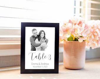 May include: A black framed table number sign with a black and white photo of a couple. The sign reads "Table 3" with the names "Emma & Andrew" and the date "November 2, 2021". A vase of pink flowers sits to the right.