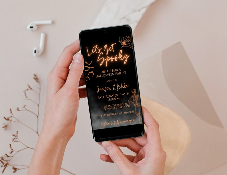 K&ouml;nnte beinhalten: Ein schwarzes Smartphone, auf dem eine Halloween-Partyeinladung mit goldener Schrift angezeigt wird, die "Let's get Spooky" lautet und Details zur Party enth&auml;lt, darunter Datum, Uhrzeit, Ort und RSVP-Informationen.