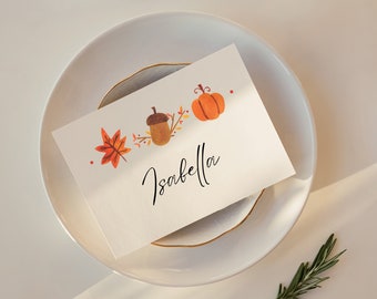May include: A white place card with a gold border and a handwritten name "Isabella". The card is decorated with watercolor illustrations of a pumpkin, acorn, and maple leaf.