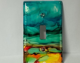 May include: A decorative light switch cover with an abstract design. The cover features a vibrant blend of colors, including shades of teal, green, yellow, orange, and red. The design has a glossy finish and includes a single switch opening and two screw holes.