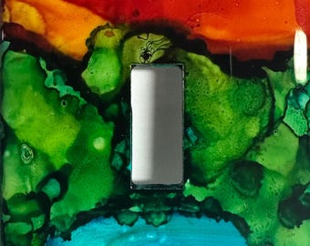 May include: A light switch cover with a colorful abstract design. The top section is red and orange, the middle section is green, and the bottom section is blue. The switch plate has two screws.