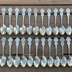 May include: A collection of forty-two silver-toned metal souvenir spoons arranged in two rows. Each spoon features a unique embossed design on the handle and a detailed engraved scene within the bowl, showcasing various landmarks and locations.