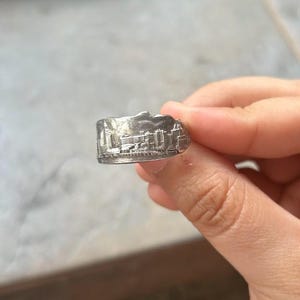 May include: A silver-toned metal ring featuring a detailed, embossed relief of a city skyline wrapping around the band. The design includes various building shapes and structures with a textured finish, creating a miniature urban landscape scene.