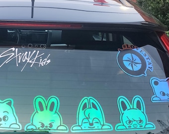 May include: A black car rear window featuring a white Stray Kids logo decal, a circular blue and black compass sticker, and a row of eight iridescent, holographic-style cartoon character stickers depicting various animal-like faces.