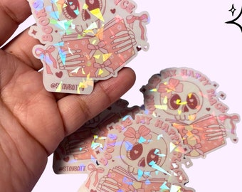 May include: Holographic stickers featuring a skeleton reading a book, with the text "Bookworm's Happy Place." The stickers have a pink and white color scheme with bow accents. The stickers are held in a hand.