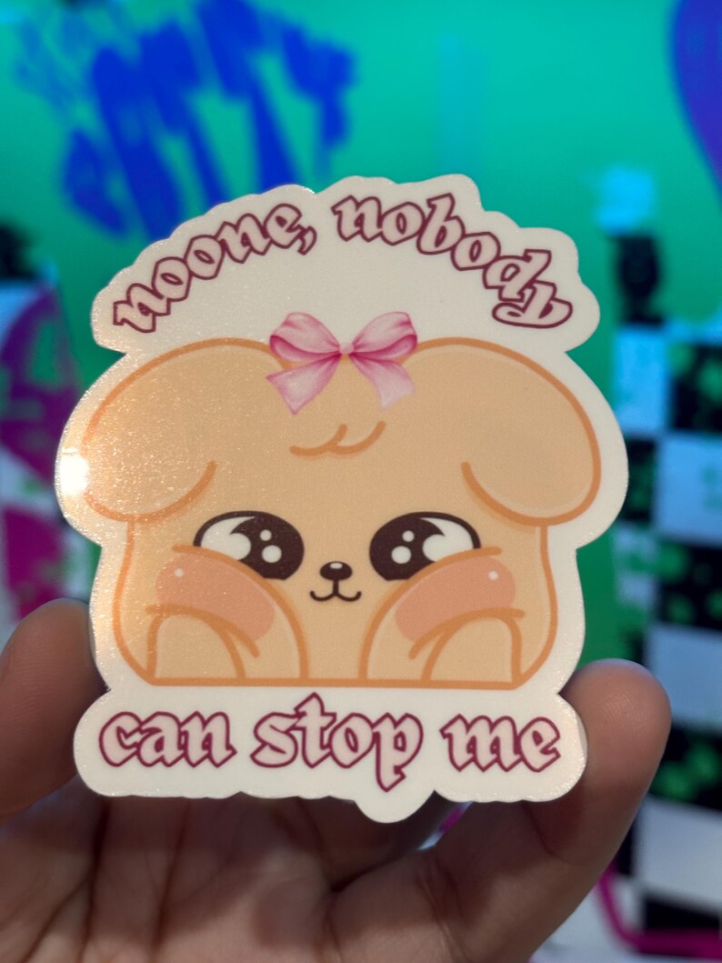 K&ouml;nnte beinhalten: Ein Aufkleber mit einem Cartoon-Hund mit gro&szlig;en Augen und rosigen Wangen. Der Hund hat eine rosa Schleife und den Text "noone, nobody" dar&uuml;ber und "can stop me" darunter. Der Aufkleber wird in einer Hand gehalten.