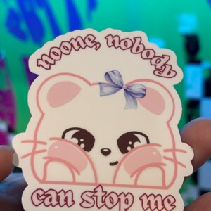 K&ouml;nnte beinhalten: Ein wei&szlig;er Aufkleber mit einem Cartoon-Maus-Design, mit rosa Wangen und einer blauen Schleife. Der Aufkleber hat den Text "noone, nobody can stop me" in einer lila Schrift. Die Maus hat gro&szlig;e braune Augen und rosa Akzente.