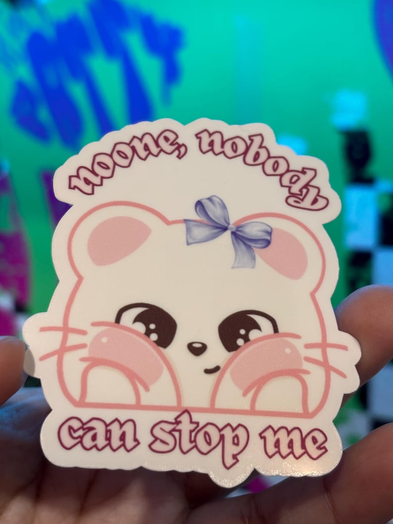 K&ouml;nnte beinhalten: Ein wei&szlig;er Aufkleber mit einem Cartoon-Maus-Design, mit rosa Wangen und einer blauen Schleife. Der Aufkleber hat den Text "noone, nobody can stop me" in einer lila Schrift. Die Maus hat gro&szlig;e braune Augen und rosa Akzente.