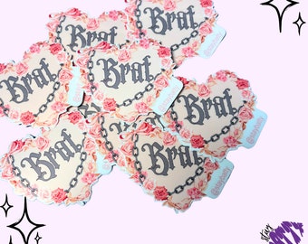 May include: A set of six heart-shaped stickers with the word "Brat" written in a gothic font. The stickers are decorated with pink roses and chains. The stickers are on a light pink background.