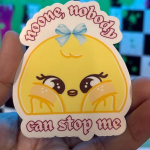K&ouml;nnte beinhalten: Ein gestanzter Aufkleber mit einer Cartoonfigur mit gelbem Gesicht, gro&szlig;en Augen und einer blauen Schleife. Der Aufkleber hat den Text "noone, nobody" &uuml;ber der Figur und "can stop me" darunter.