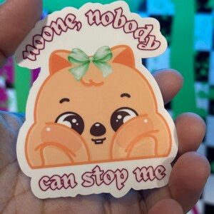 K&ouml;nnte beinhalten: Ein Aufkleber mit einem Cartoon-Quokka mit gro&szlig;en, ausdrucksstarken Augen und einer gr&uuml;nen Schleife. Der Aufkleber hat den Text "noone, nobody can stop me" in einer kursiven Schrift. Das Quokka ist orange und wei&szlig;.
