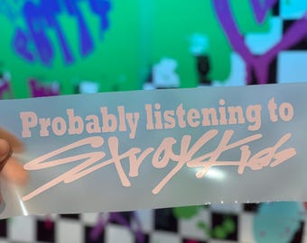 May include: A clear sticker with the text "Probably listening to Stray Kids" in a light pink color. The background is a mix of colors and patterns, including a checkered design and graffiti art.