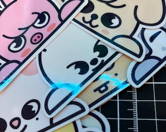 May include: A stack of colorful cartoon animal stickers. The stickers feature a pink bunny, a tan dog, a white bear, and a yellow duck. Each sticker has a black outline and a unique facial expression. The stickers are on a black cutting mat.
