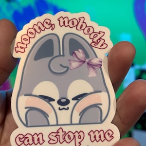 K&ouml;nnte beinhalten: Aufkleber mit einem Cartoon-Tier-Design. Das Tier ist hellgrau mit rosa Wangen und einer lila Schleife. Der Aufkleber hat den Text "noone, nobody can stop me" in rosa.